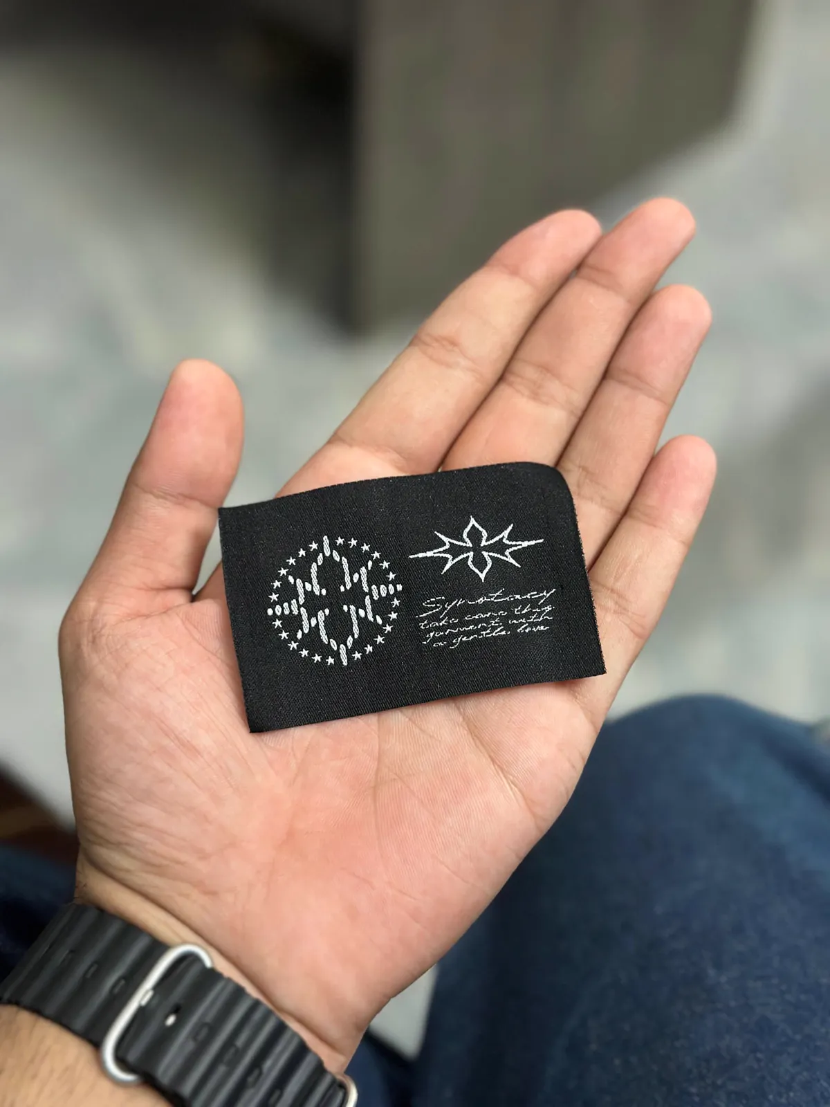 A woven black Synotacy label held in an open palm.