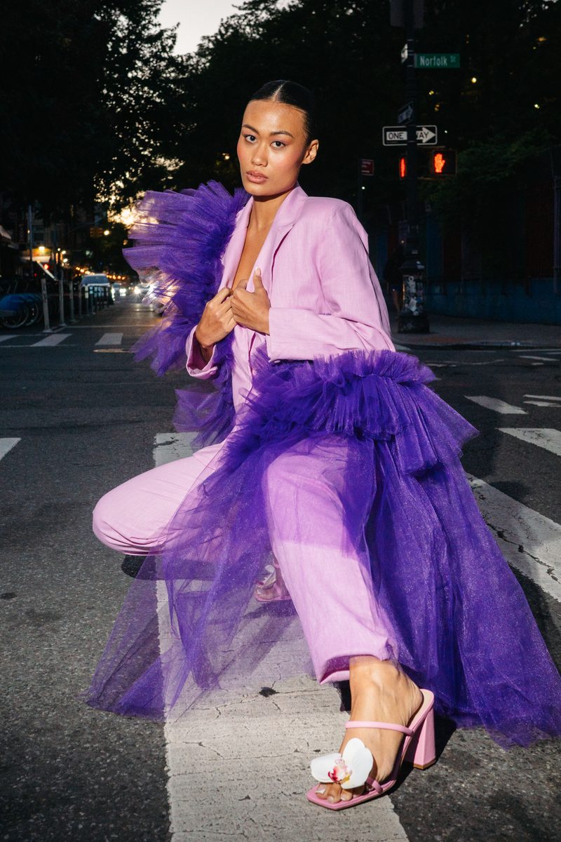 A model in a lilac suit and violet tulle coat crouches on a New York crosswalk at dusk.