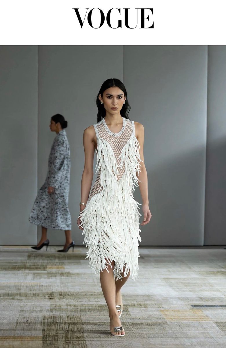 Vogue: a model in a white fringed dress walks a runway as a second model passes behind.