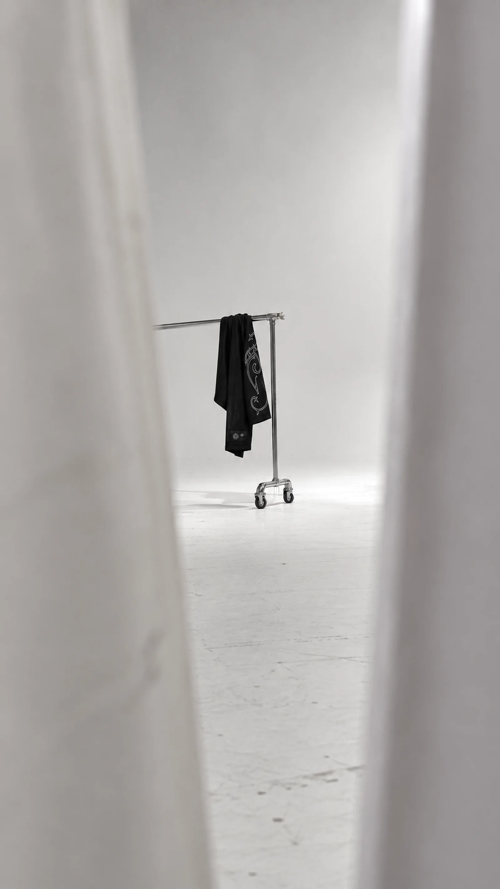A single black Synotacy garment hangs on a steel rail in a white studio, seen between pale curtains.